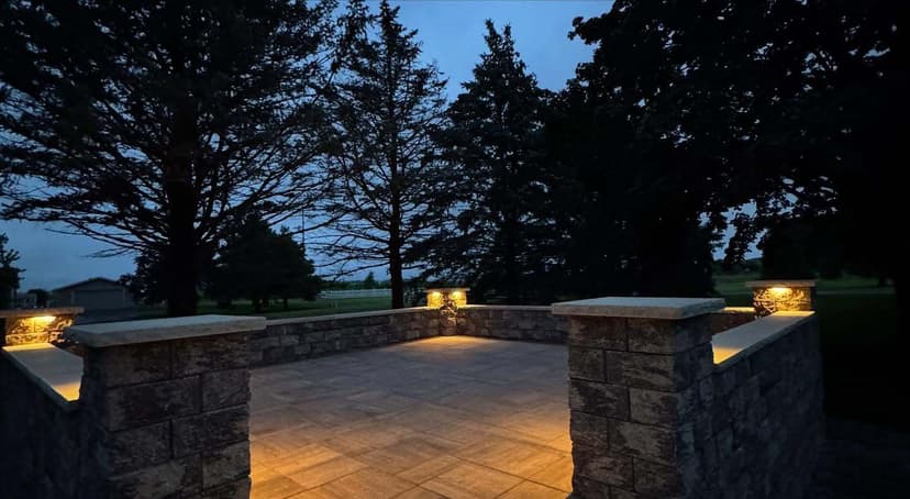 Evening view of a stone patio illuminated by lights, surrounded by tall trees.