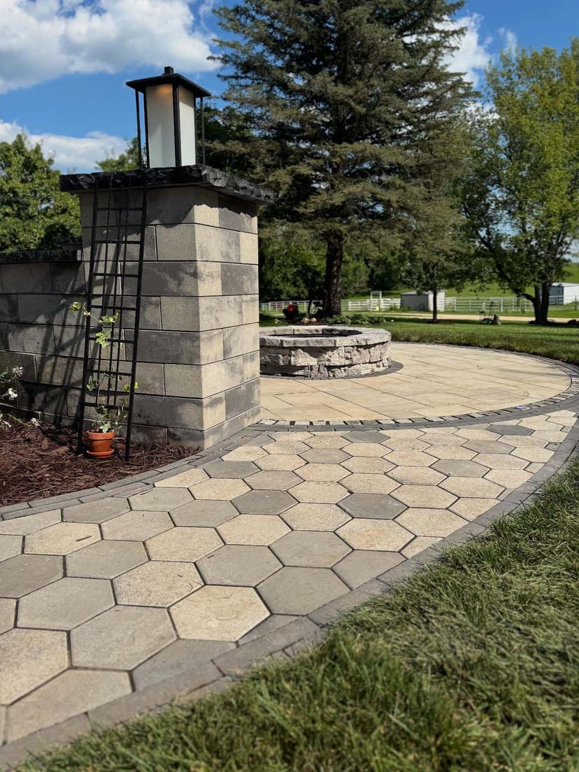 Hexagonal paver pathway winding past a stone fireplace and lantern in a landscaped yard.